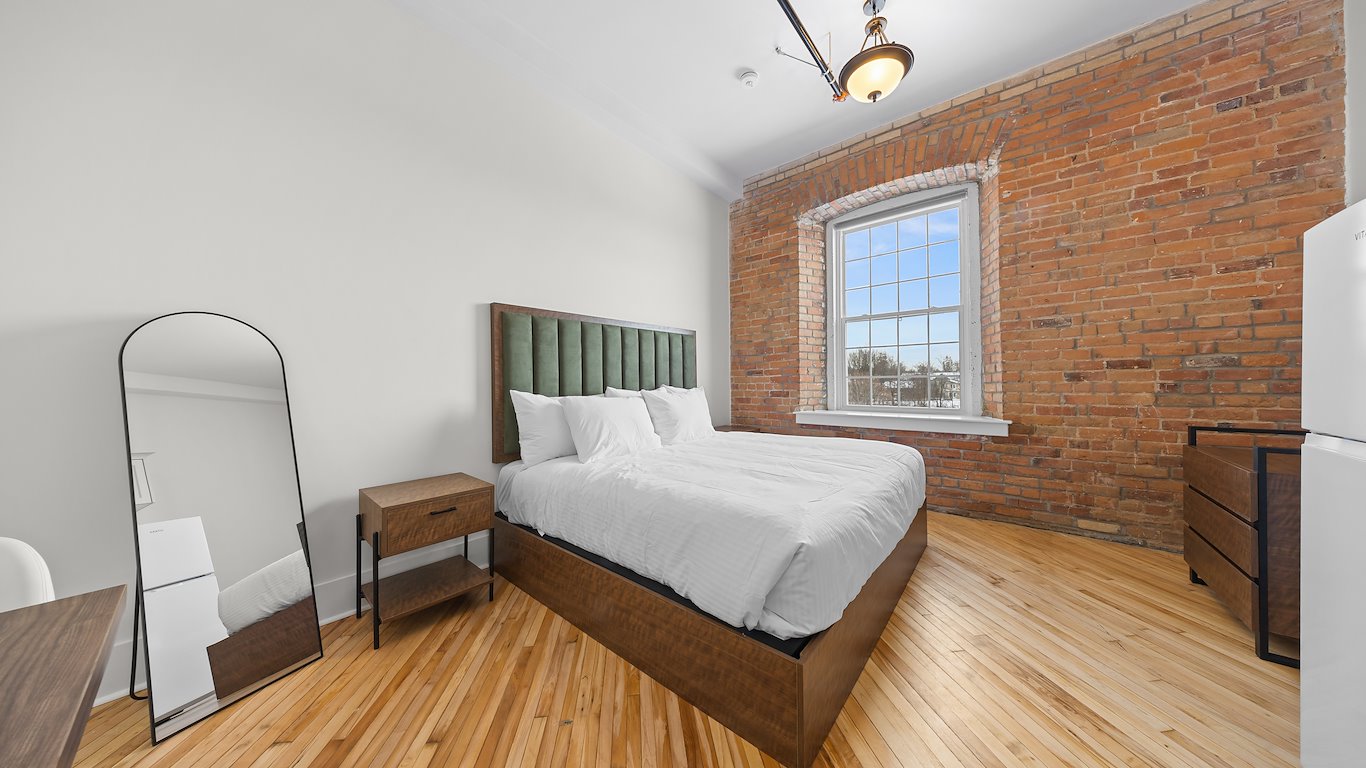 A modern bedroom with a brick accent wall, large window, wood floors, double bed with green headboard, nightstand, tall mirror, dresser, and overhead light fixture.
