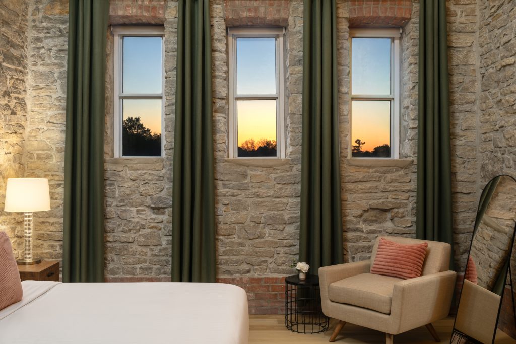 A cozy bedroom with stone walls, three tall windows showing a sunset, a beige armchair with a pillow, a floor lamp, and a small round table with a plant.