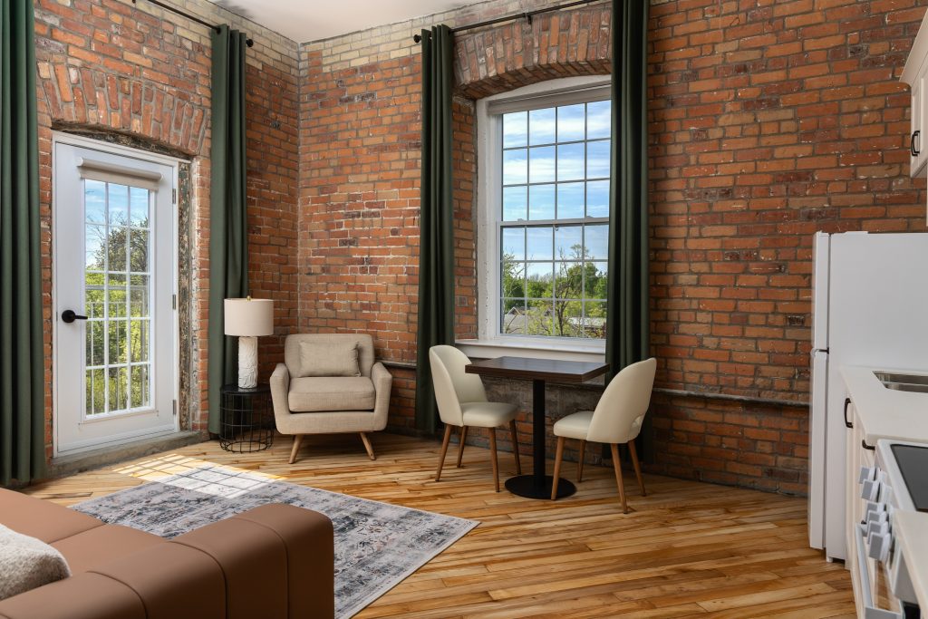 A modern loft with exposed brick walls, a large window, wood floors, a small table with two chairs, an armchair, and a lamp near a glass door leading outside.