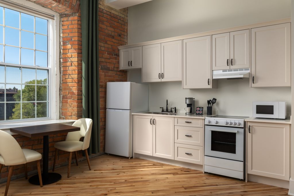 A small kitchen with white cabinets, a stove, refrigerator, microwave, coffee maker, and a dining table with two chairs near a large window with brick walls.