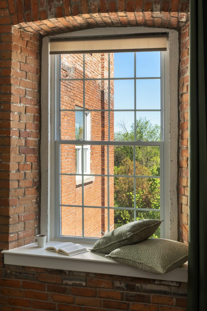 A window with a grid frame set in a brick wall, showing an adjacent brick building, green trees, a book, pillows, and a white mug on the windowsill.