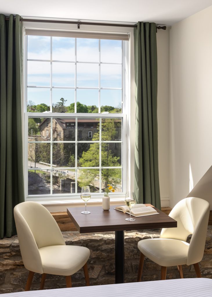 A small table with two chairs, wine glasses, a book, and a flower vase is set by a window with green curtains, overlooking a building and trees outside.