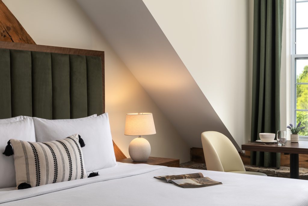 A neatly made bed with white linens, a decorative pillow, a bedside lamp, and a small table with a chair by a window with green curtains.