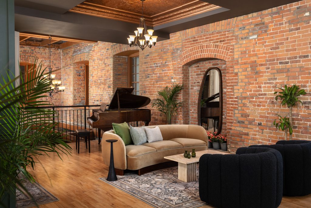 A modern living room with exposed brick walls, a beige sofa, black armchairs, a coffee table, indoor plants, and a grand piano in the background.