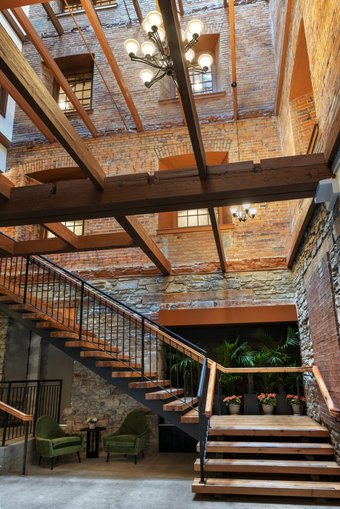 Exposed brick and wood beams frame a modern staircase with metal railings in an indoor courtyard featuring green chairs, potted plants, and large windows.