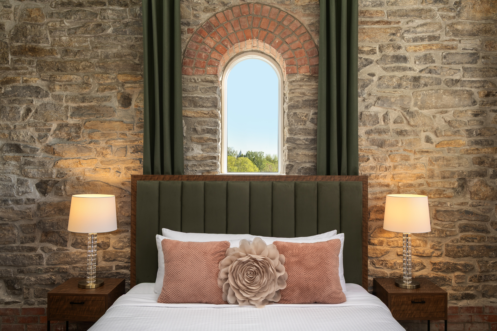 A bed with a green upholstered headboard, pink pillows, and a floral cushion sits between two lamps and nightstands against a stone wall with an arched window.